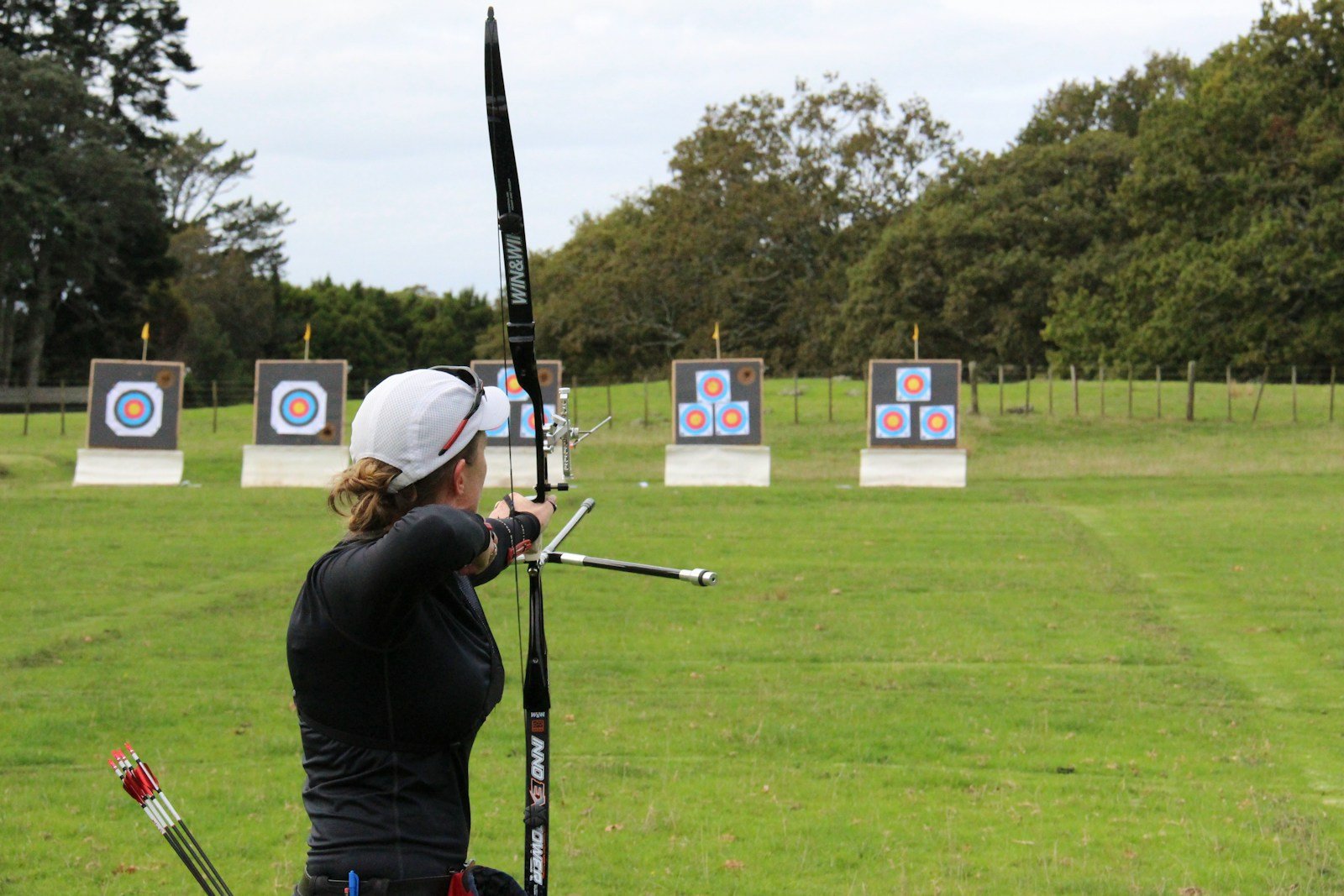 Woman archer at full draw seen from behind on an archery field, bow arm reaching toward the targets