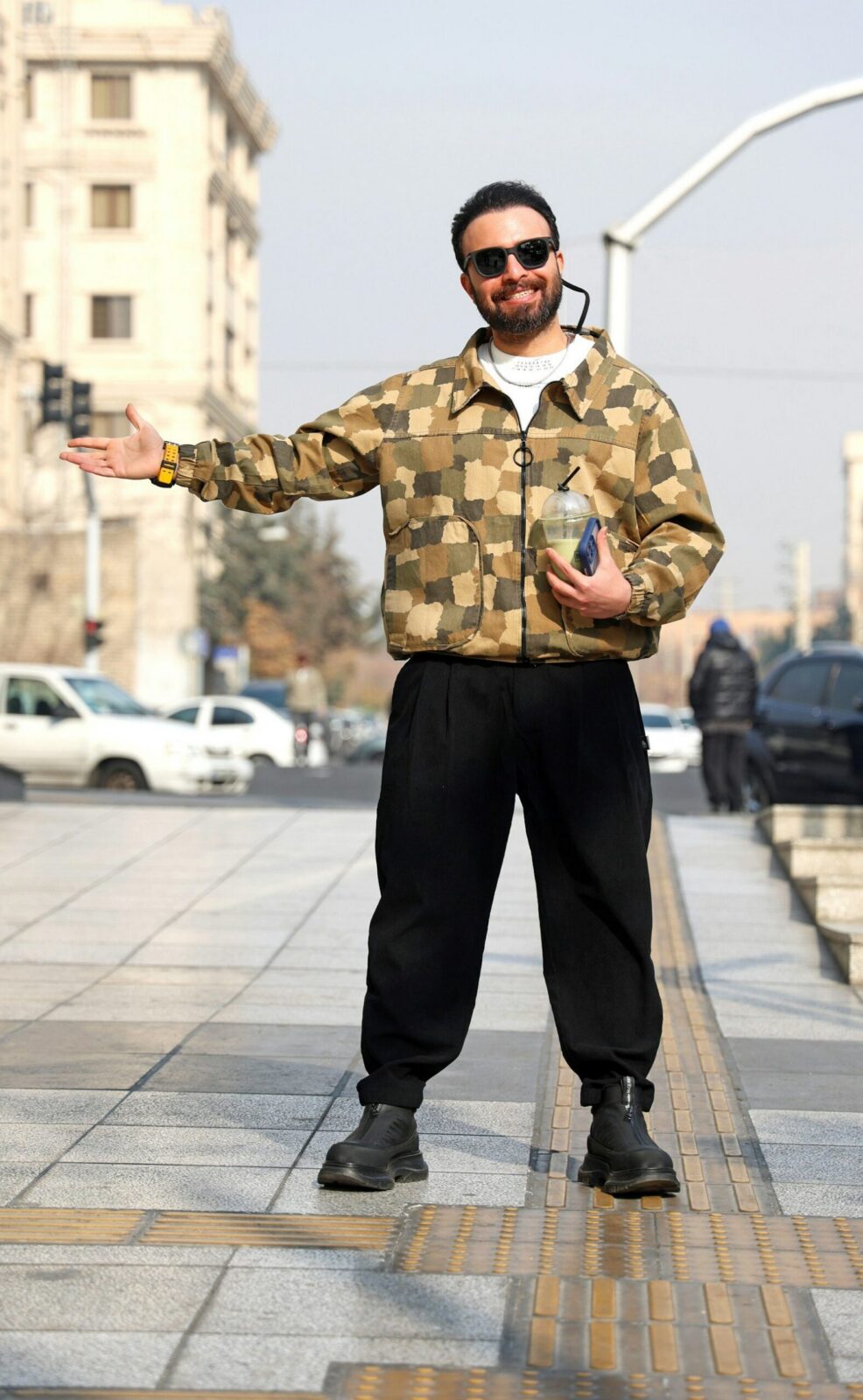 Man in a camouflage jacket and black trousers standing on a street with his arms out