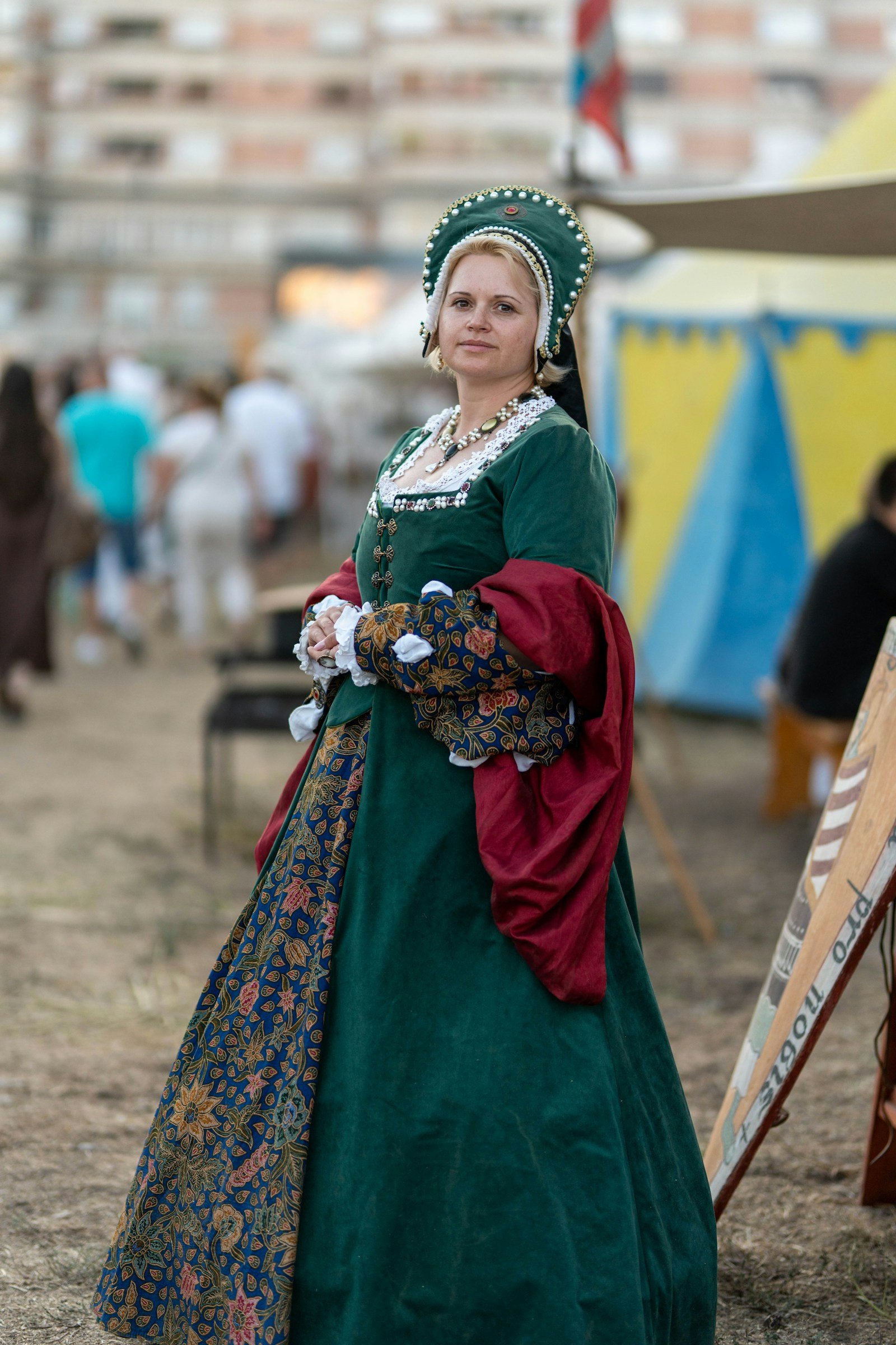 Woman in a long green medieval dress with elaborate headwear and red cloth over one arm