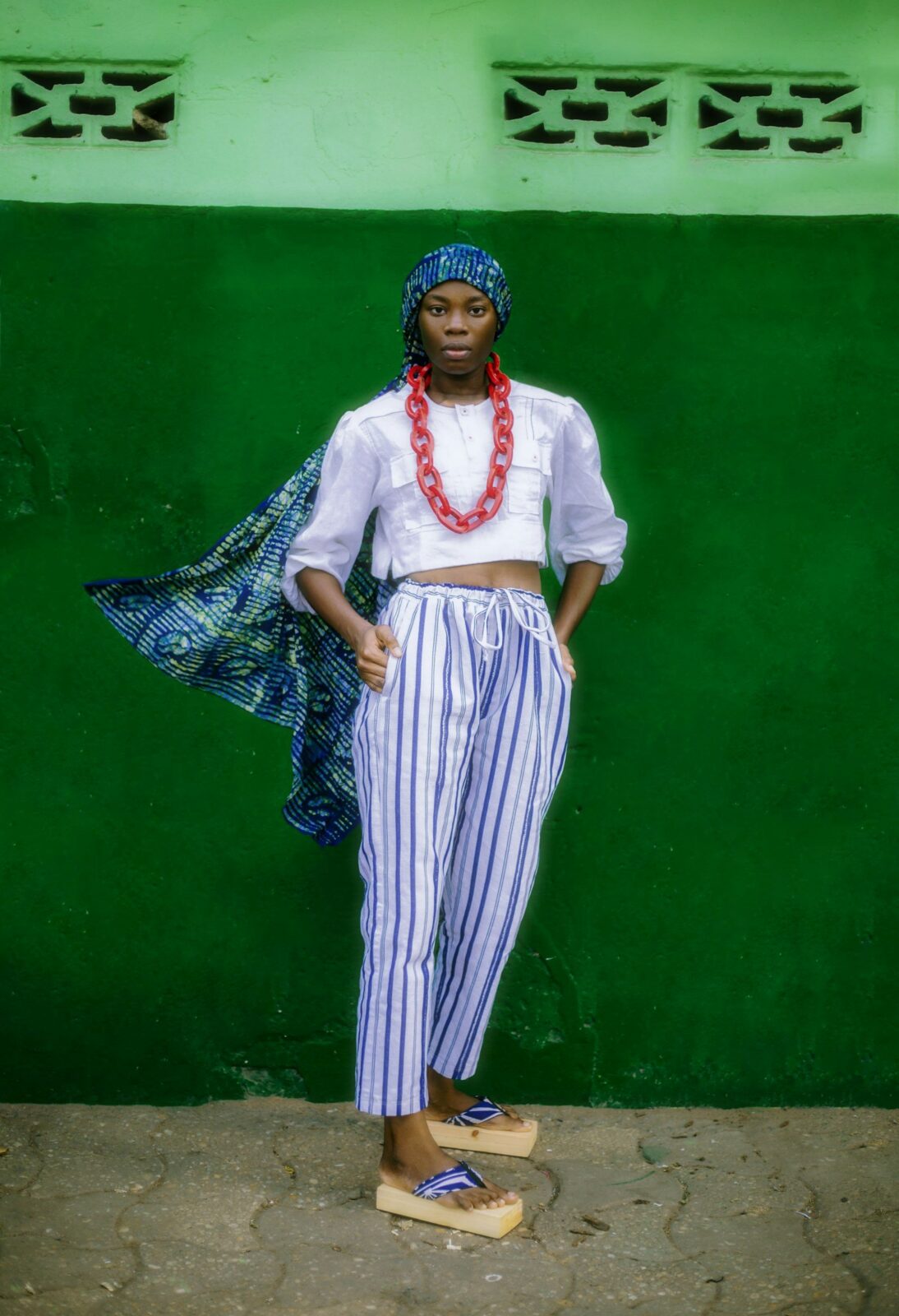 Woman in striped trousers, a white shirt and a headscarf against a green wall