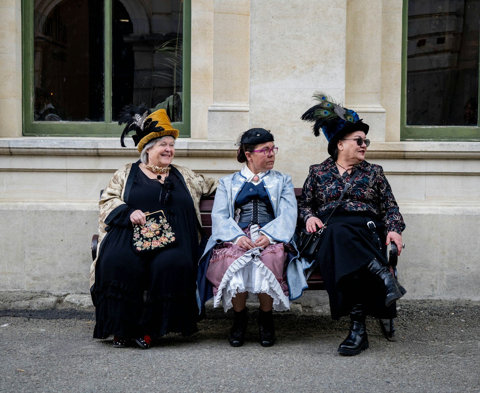 Three women seated on a bench in Victorian-era hats and dark dresses
