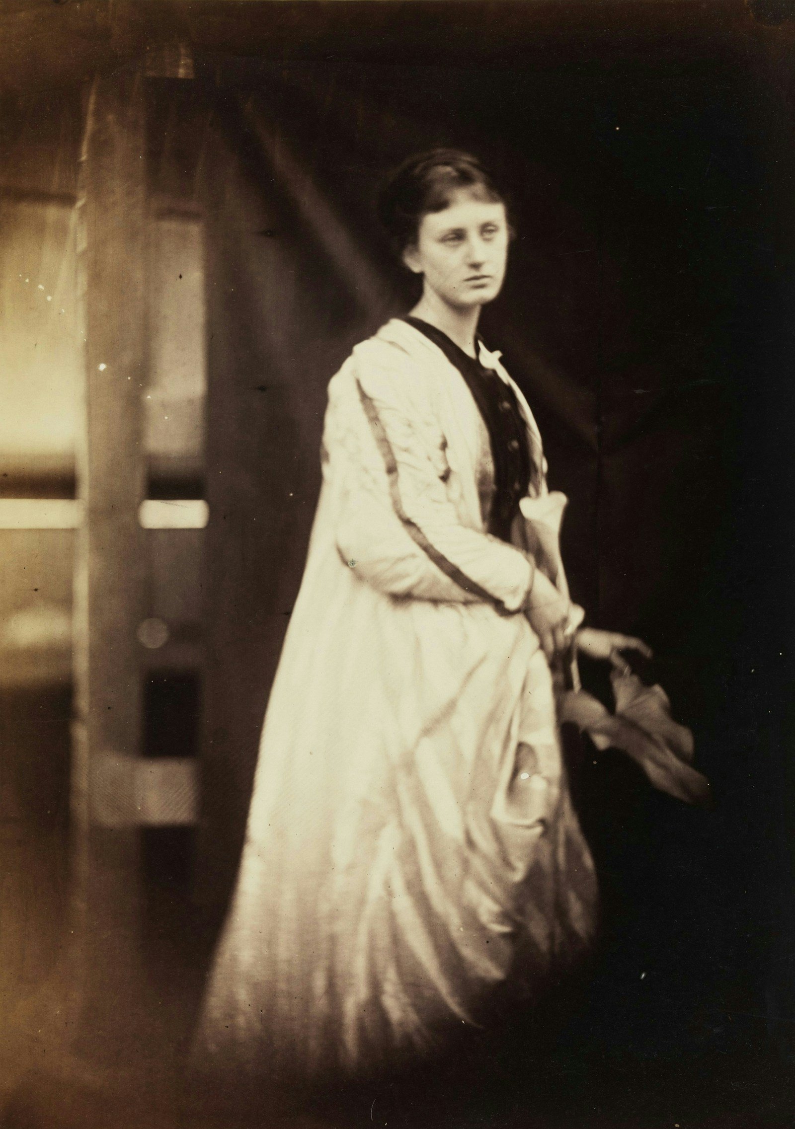 Victorian woman standing in a long pale gown against a dark background