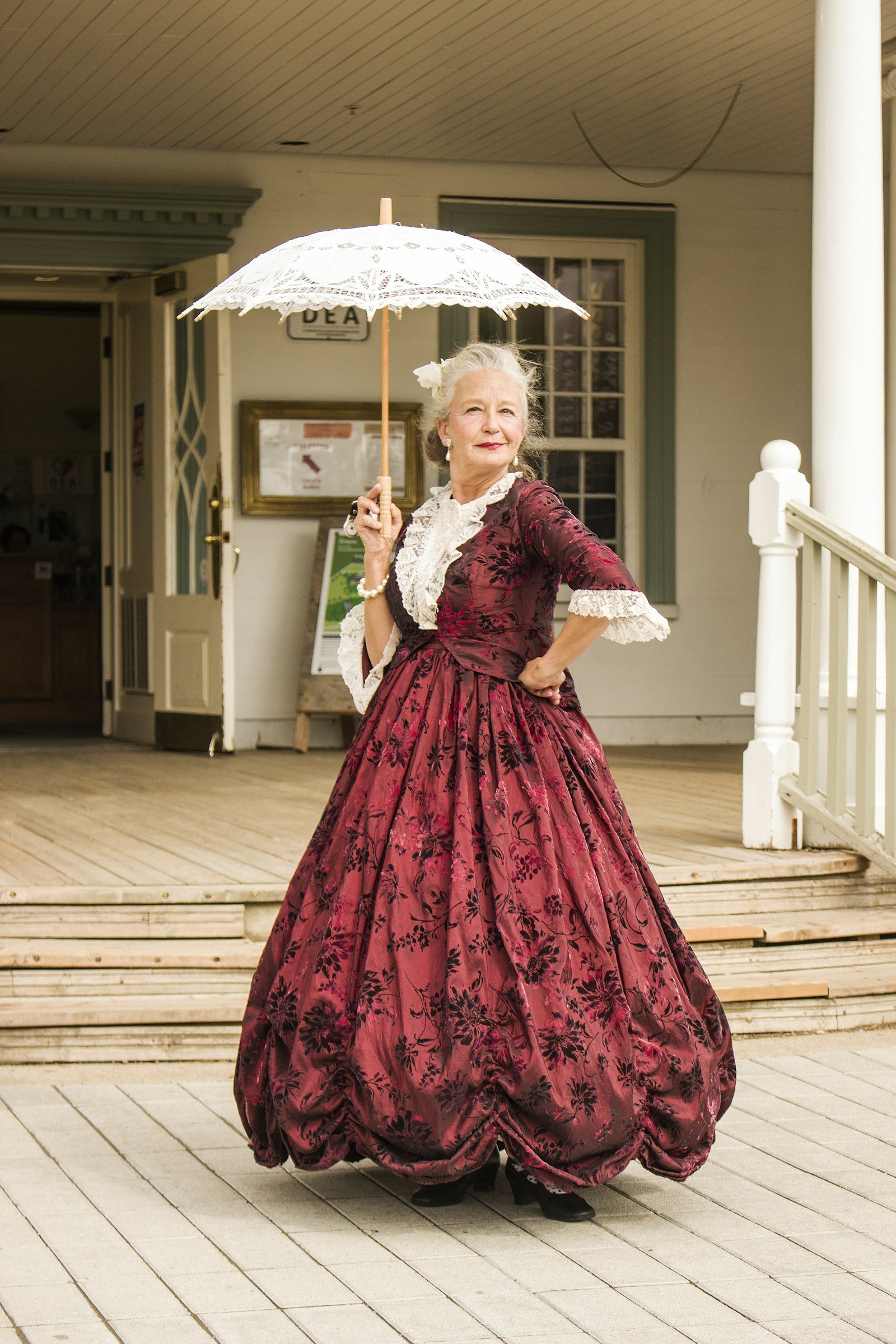 Woman in a vintage red floral dress holding a white lace parasol in front of an old building