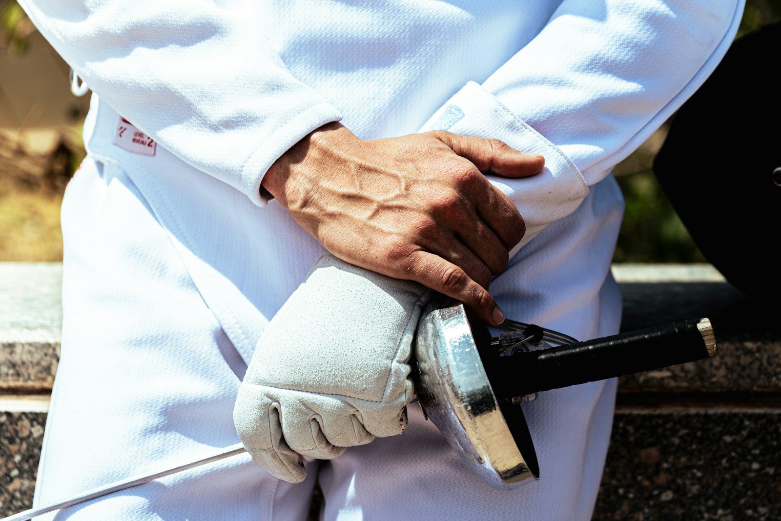 Fencer in white with a gloved hand holding a foil by its guard behind the back