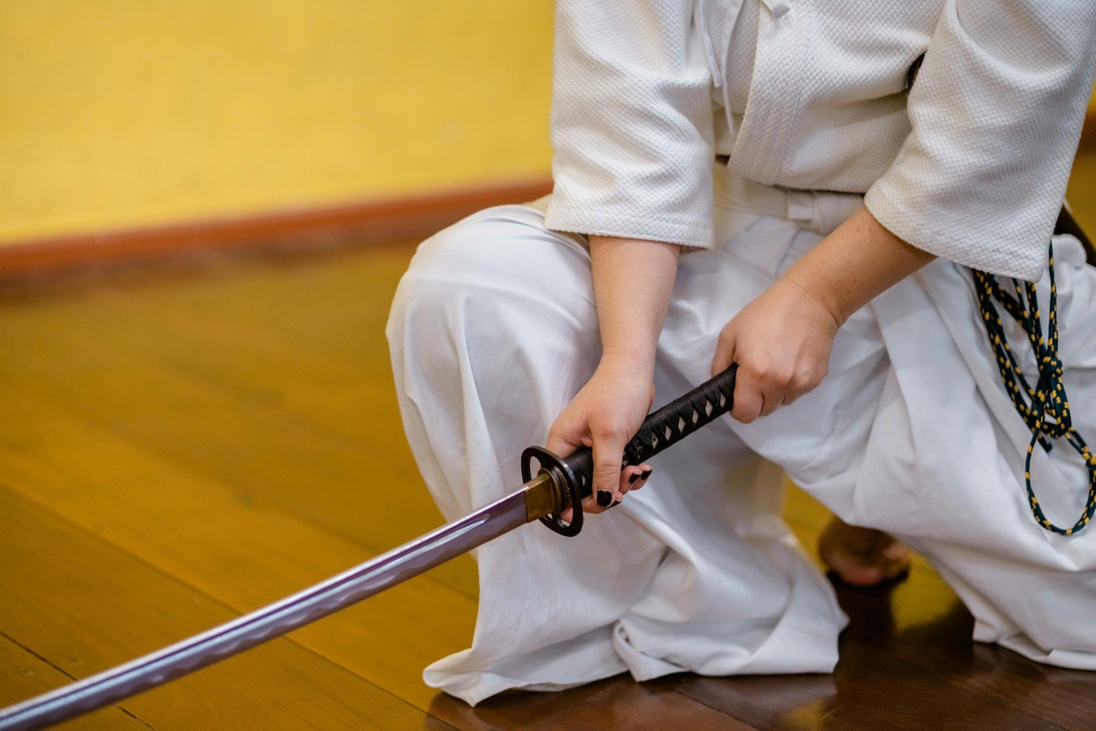 Person in a white training uniform kneeling with a katana, one hand on the hilt